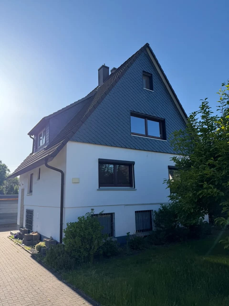 Freistehendes Einfamilienhaus mit Satteldach, schiefergedeckter Giebelseite, weiß verputztem Erdgeschoss und Schleppgaube, aufgenommen von der Auffahrt