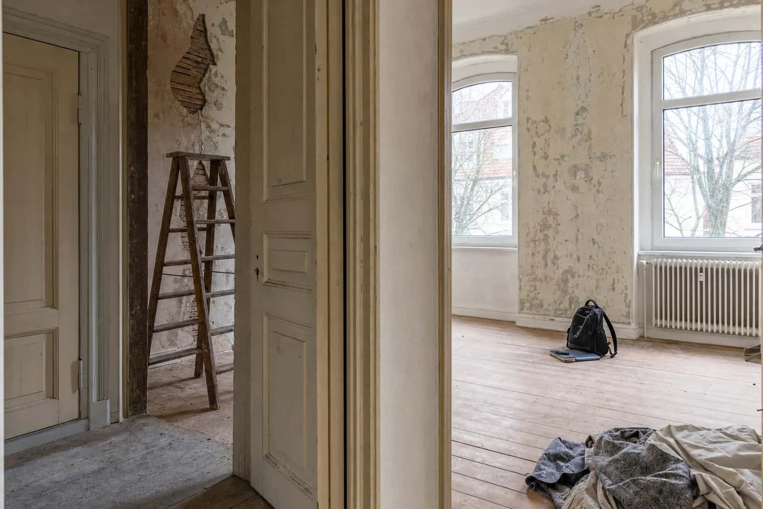Zwei helle, renovierungsbedürftige Räume mit abblätternder Wandfarbe, Holzboden, Leiter, Rucksack und Heizkörper